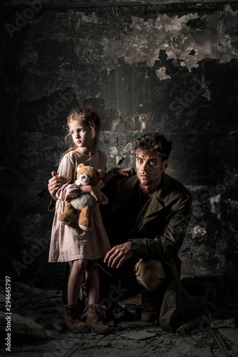 handsome man sitting near upset kid with teddy bear in dirty room, post apocalyptic concept © LIGHTFIELD STUDIOS