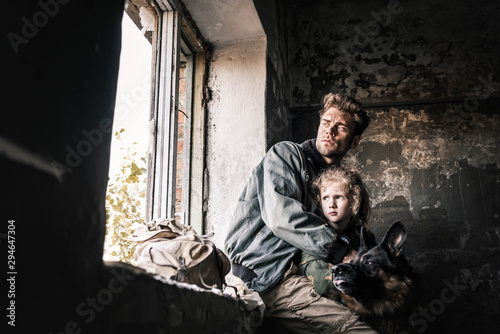 man hugging dirty kid near german shepherd dog in abandoned building, post apocalyptic concept