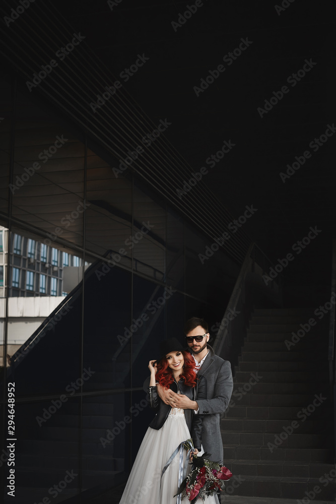 Fototapeta premium Beautiful couple of lovers hugging outdoors during wedding photoshoot. Slim young woman with red hair in modish leather jacket and wedding dress and handsome bearded man in the coat.