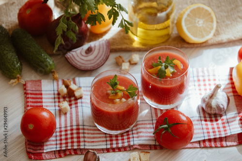 Tomato soup gazpacho in glass with herbs and ingredients on the kitchen. A beautiful summer soup spilled in glasses surrounded by juicy vegetables