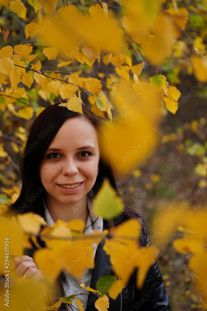 Portrait of a beautiful smiling woman is standing in the autumn forest between the branches of trees with yellow foliage.