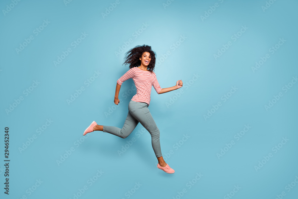 Full length body size photo of cute nice charming cheerful sweet curly wavy girl running in trousers pants striped t-shirt footwear smiling toothily aspiring forward isolated over blue pastel color