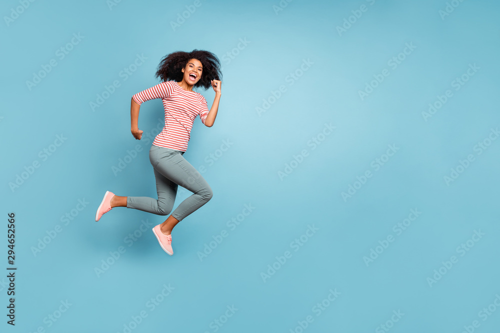 Full length body size profile side photo of cute running jumping girl wearing striped t-shirt trousers pants footwear rejoicing isolated over pastel blue color background