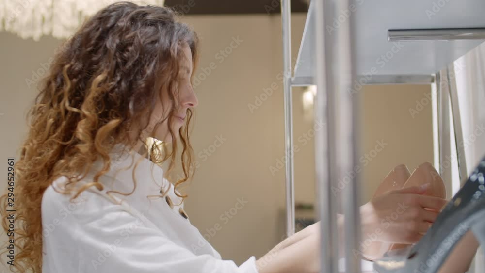 Close up of Caucasian woman with long curly hair standing at rack in shoe shop and choosing patent leather shoes Close up of Caucasian woman with long curly hair standing at rack in shoe shop and choosing patent leather shoes