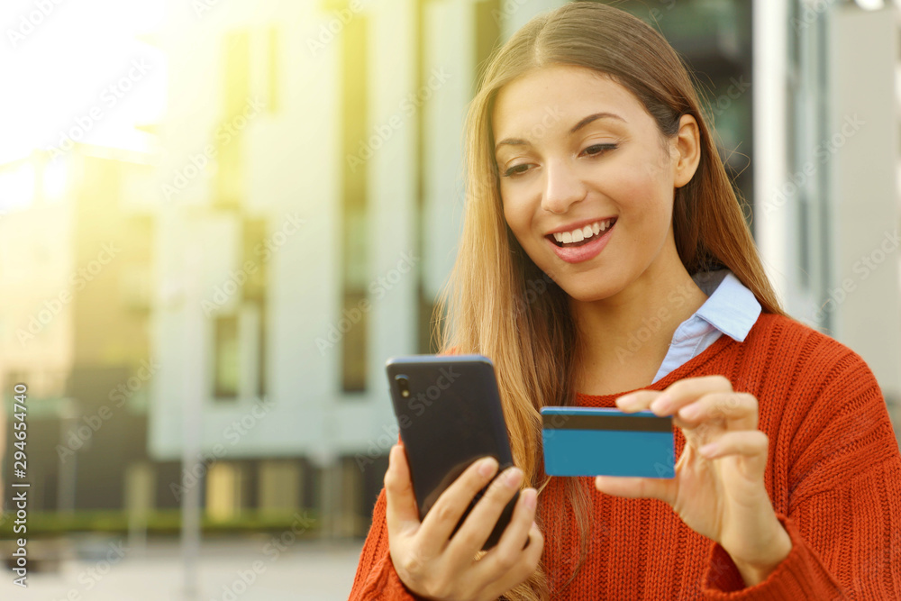 Happy smiling woman wearing sweater paying online holding with smart phone and credit card outdoors in autumn season. Copy space. Happy smiling woman wearing sweater paying online holding with smart phone and credit card outdoors in autumn season. Copy space.