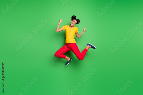 Full size photo of pretty dark skin lady jumping high celebrating exciting victory raising fists wear casual yellow t-shirt red pants isolated green background © deagreez