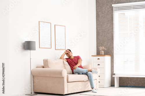 Young man with air conditioner remote control suffering from heat on sofa at home