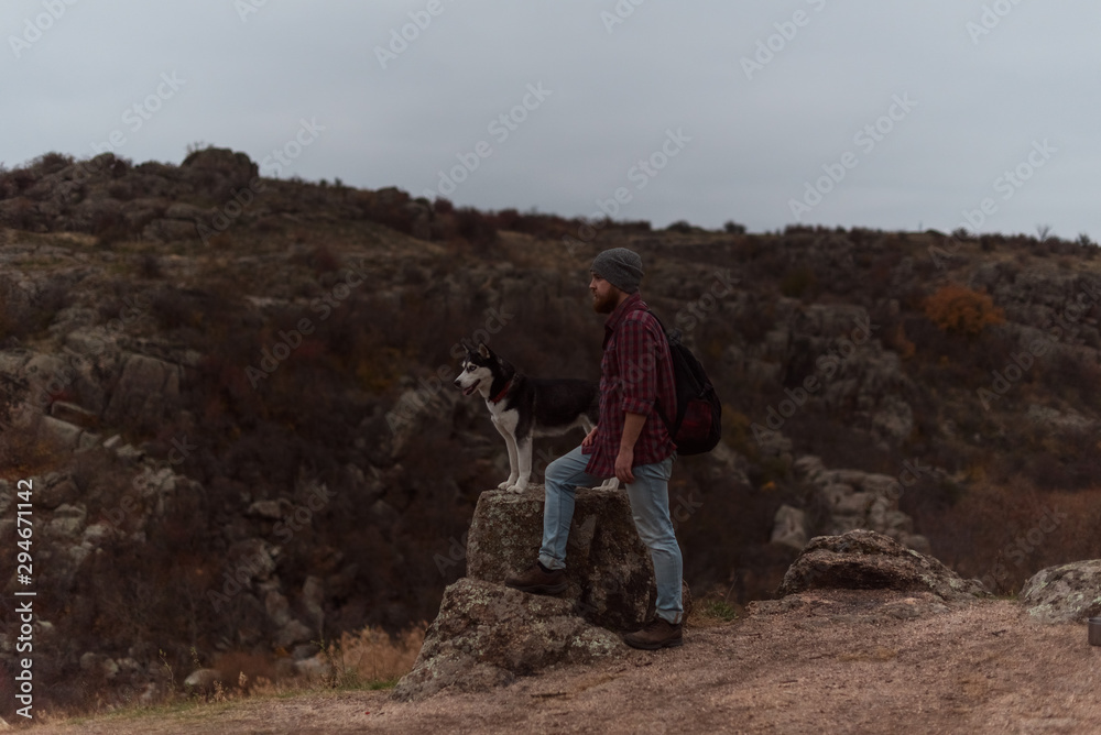 Bearded tourist in plaid shirt with a beard and backpack and a husky dog on a stone ​​on the background of a gorge and stones