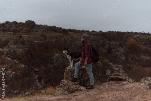 Bearded tourist in plaid shirt with a beard and backpack and a husky dog on a stone ​​on the background of a gorge and stones © iwavephoto