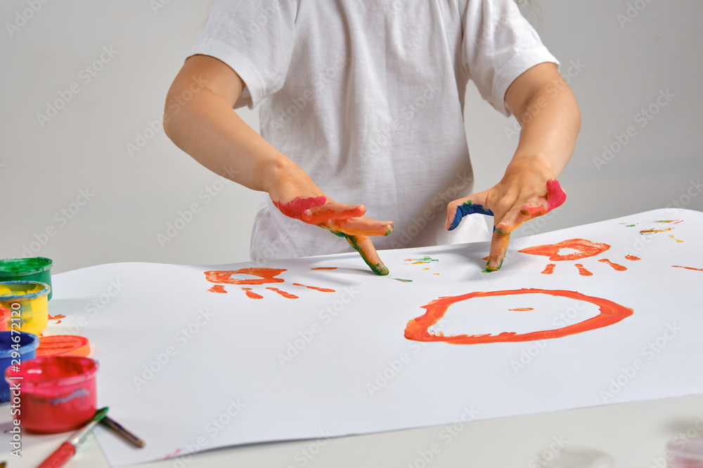 Little artist in white t-shirt standing at table with whatman and colorful paints, painting on it with her hands. Isolated on white. Close-up. Little artist in white t-shirt standing at table with whatman and colorful paints, painting on it with her hands. Isolated on white. Close-up.