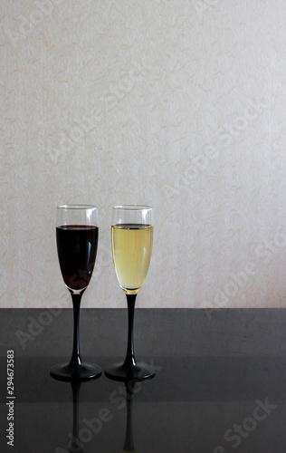 Two wineglasses with white and red wine on black glass table. Gray blurred wall behind. Background with dark and light liquids in goblets and place for text.