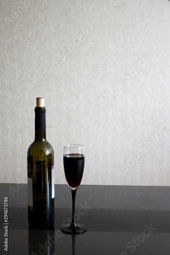 Wineglasses with red wine and bottle on black glass table. Gray blurred wall behind. Background with light liquid in goblet and place for text.
