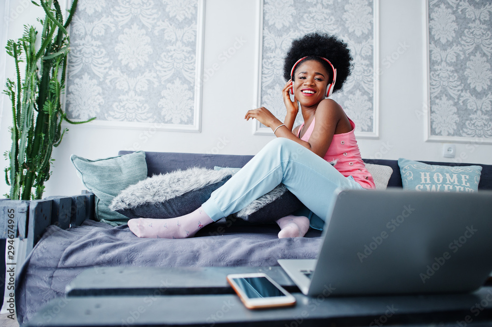 Young african american woman sitting on the couch while working on laptop and listen music on earphones. Young african american woman sitting on the couch while working on laptop and listen music on earphones.