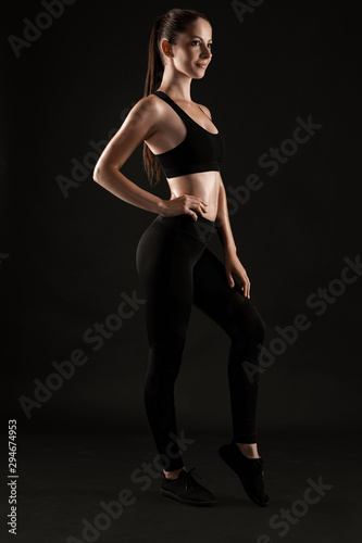 Brunette woman in black leggings, top and sneakers is posing against a black background. Fitness, gym, healthy lifestyle concept. Full length.