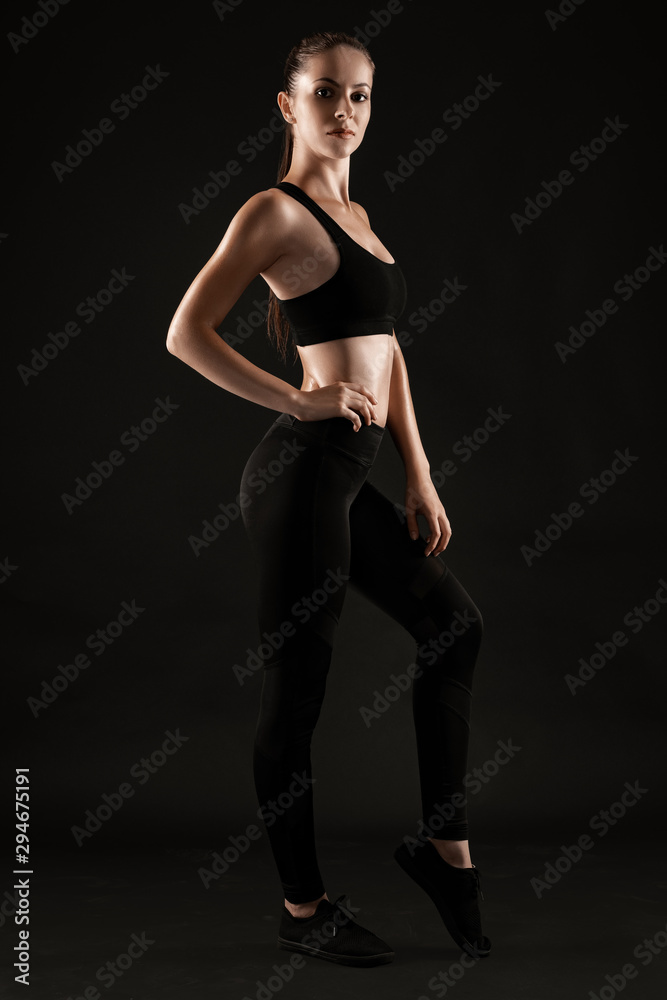 Brunette woman in black leggings, top and sneakers is posing against a black background. Fitness, gym, healthy lifestyle concept. Full length.