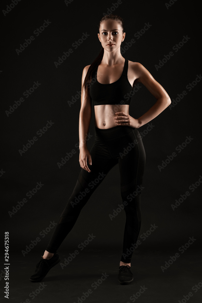 Brunette woman in black leggings, top and sneakers is posing against a black background. Fitness, gym, healthy lifestyle concept. Full length. Brunette woman in black leggings, top and sneakers is posing against a black background. Fitness, gym, healthy lifestyle concept. Full length.