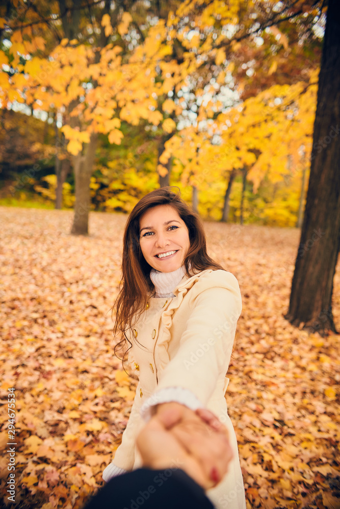Portrait of cheerful young woman holding boyfriend by hand in autumn park. Smiling lady walking in autumn wood with her friend. Season, friendship, dating Portrait of cheerful young woman holding boyfriend by hand in autumn park. Smiling lady walking in autumn wood with her friend. Season, friendship, dating