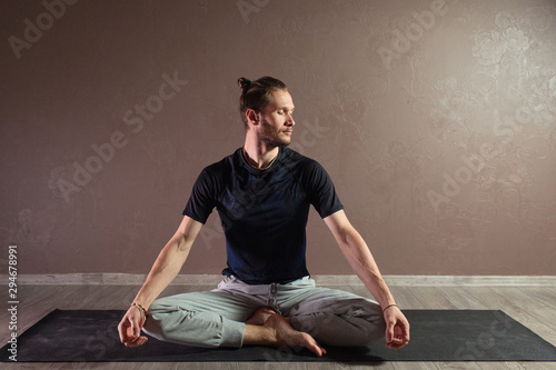 Young sporty man practicing yoga, meditating in Half Lotus pose, working out, wearing sportswear, indoor full length, gray yoga studio