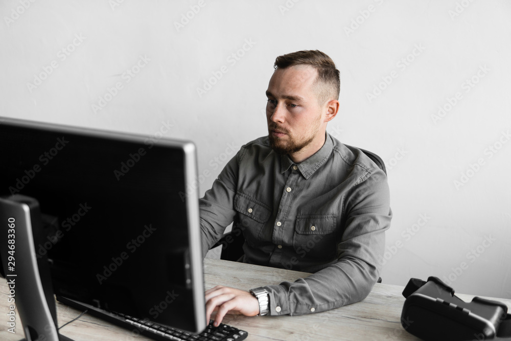 Young businessman or student in a shirt sitting against monitor of computer. Working on a pc at a table in the office with a thoughtful expression. Young businessman working on his laptop in office. Young businessman or student in a shirt sitting against monitor of computer. Working on a pc at a table in the office with a thoughtful expression. Young businessman working on his laptop in office.