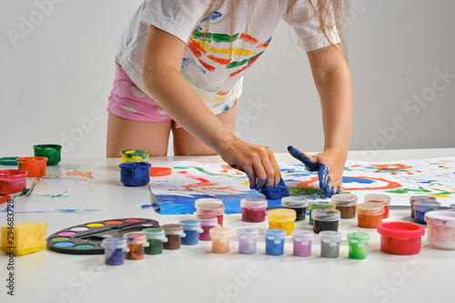 Little artist in white t-shirt standing at table with whatman and colorful paints, painting on it with her hands. Isolated on white. Close-up.