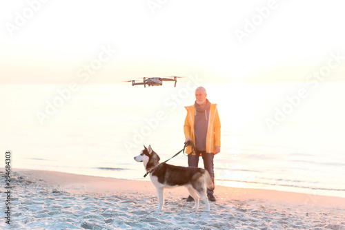 Old man in a yellow raincoat with husky dog on leash shooting on drone the beach