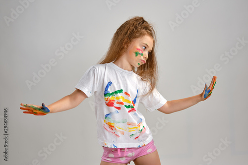 Little girl in white painted t-shirt is posing standing isolated on white and gesticulating with her colored in different paints palms and face. Art studio. Close-up.