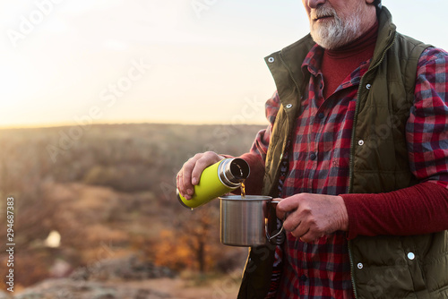 Eldery tourist man with gray hair and beard in plaid shirt drinking tea from a thermos to an iron mug against the background of the gorge and stones
