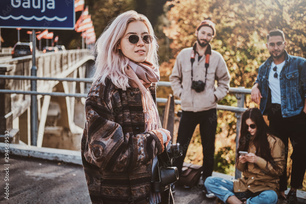 Attractive blond girl with photo camera is enjoying autumn walk with her friends.