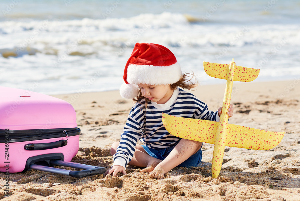 Child traveler. Little girl in Santa hat playing with toy airplane on sunny tropical beach. pulling a suitcase luggage. Christmas and new year family vacation, travel concept Child traveler. Little girl in Santa hat playing with toy airplane on sunny tropical beach. pulling a suitcase luggage. Christmas and new year family vacation, travel concept
