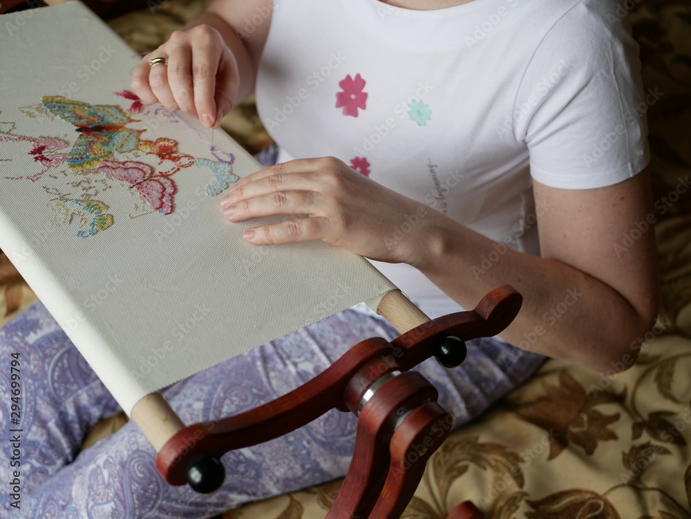 Fototapeta premium a young woman with beautiful hair in a white t-shirt embroiders colorful butterflies on a special wooden machine. Multicolored floss on a white canvas.