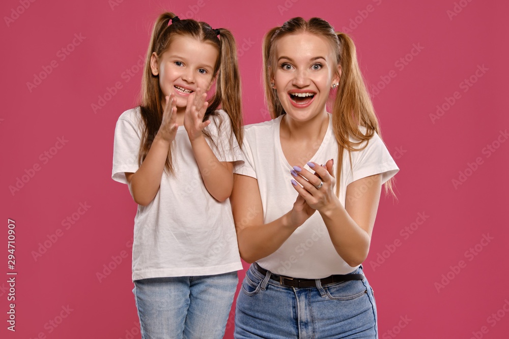 Mom and daughter with a funny ponytails, dressed in white t-shirts and blue denim jeans are posing against a pink studio background. Close-up shot. Mom and daughter with a funny ponytails, dressed in white t-shirts and blue denim jeans are posing against a pink studio background. Close-up shot.