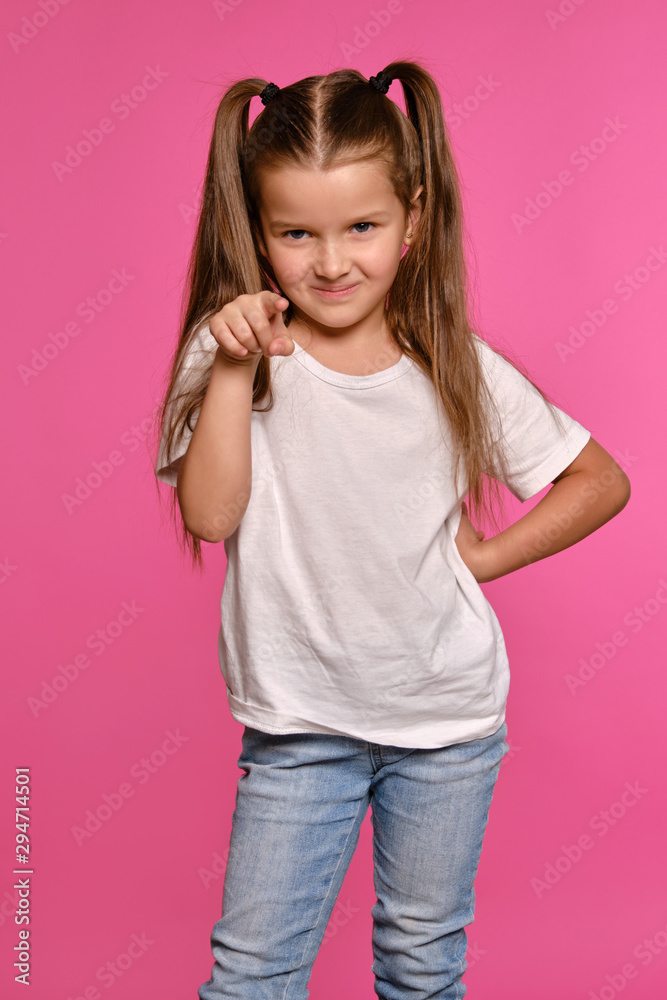 Little girl with ponytails, dressed in white t-shirt and blue jeans is posing against a pink studio background. Close-up shot. Sincere emotions. Little girl with ponytails, dressed in white t-shirt and blue jeans is posing against a pink studio background. Close-up shot. Sincere emotions.