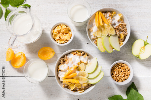 Two bowls with granola, fruit, yogurt and two glasses with milk on a white wooden background. Healthy breakfast cereal top view
