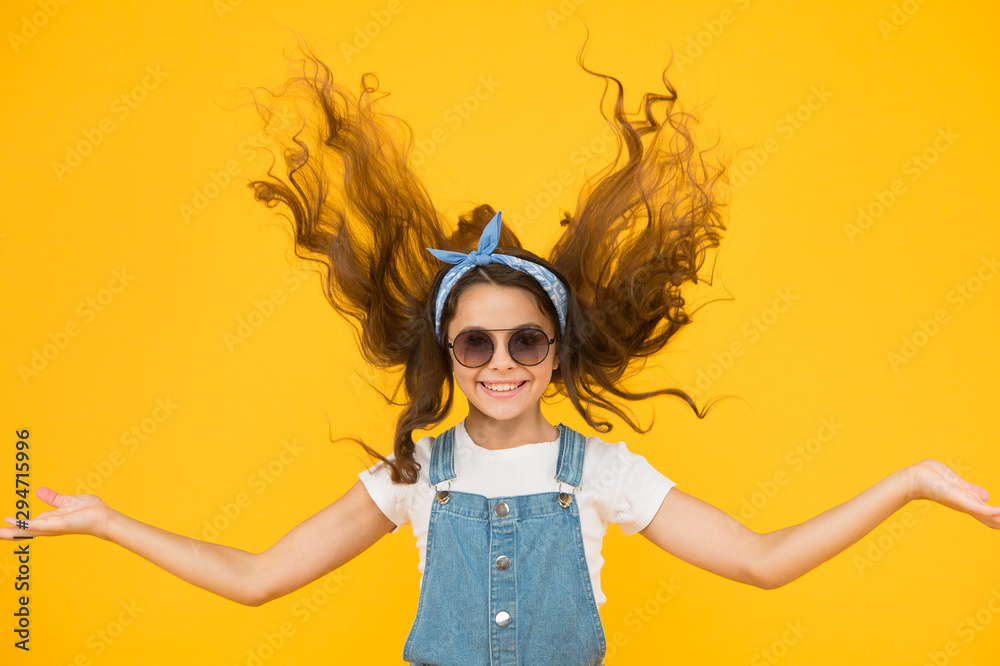 Feeling good fabulous hair. Adorable girl with curly hair waving on yellow background. Little hair model with fashion look. Small cute girl with long brunette hair. Hairdressers or beauty salon