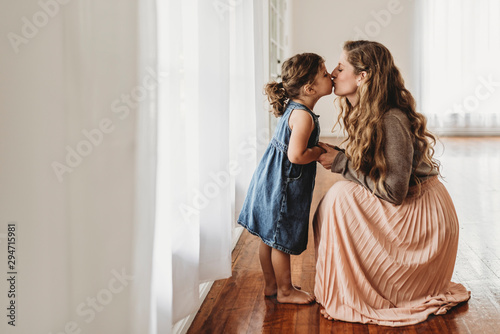 Mother bending down to kiss young daughter in natural light studio photo
