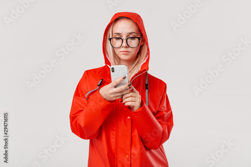 Blond woman in a red raincoat with a hood checking smartphone for notifications isolated over white background. Going for a walk on a rainy cold day photo