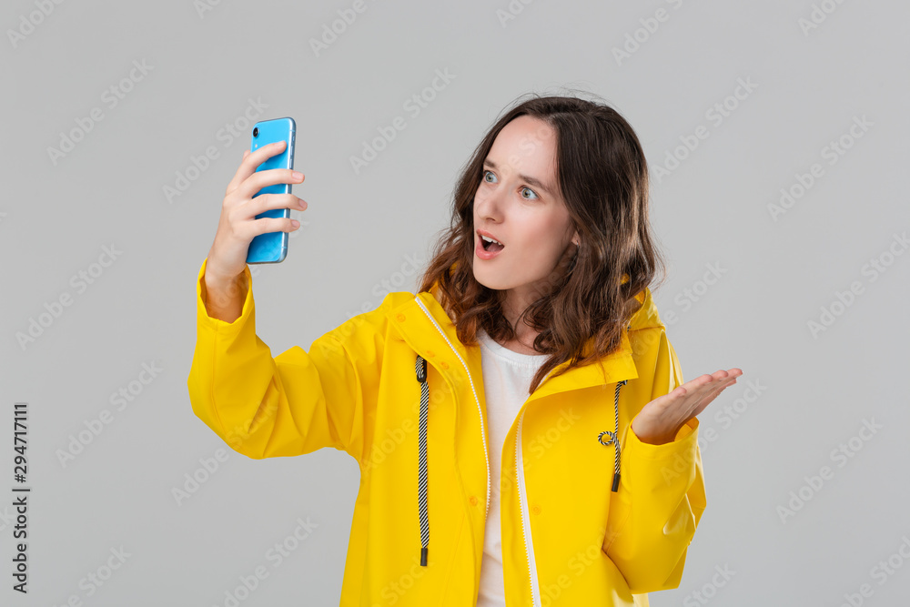 Pretty brunette woman in a yellow raincoat video chatting on the smartphone isolated over grey background. Concept of communication. Pretty brunette woman in a yellow raincoat video chatting on the smartphone isolated over grey background. Concept of communication.