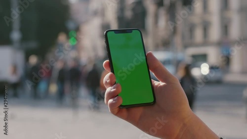 Man's hand showing vertical smartphone with mock-up chroma key greenscreen on outdoor background. Young person with modern iphone in busy city street close-up.