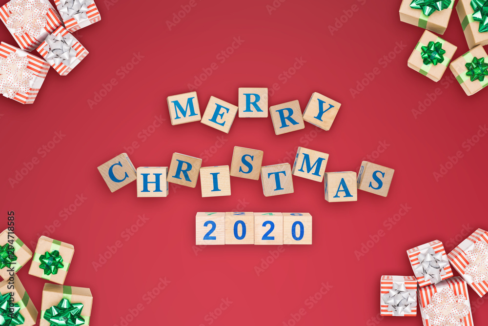 Christmas design, text inscription Merry Christmas 2020 laid out of wooden cubes on red background, many gift boxes are located around perimeter