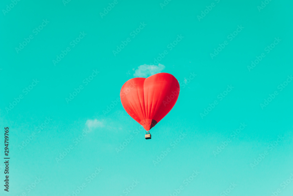 Obraz premium Red hot air balloon in the shape of a heart against the blue sky and some light clouds.