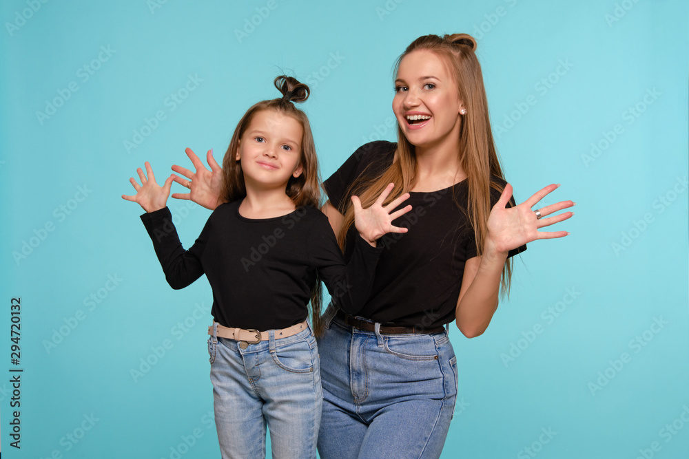 Mom and daughter with a funny hairstyles, dressed in black shirts and blue denim jeans are posing against a blue studio background. Close-up shot. Mom and daughter with a funny hairstyles, dressed in black shirts and blue denim jeans are posing against a blue studio background. Close-up shot.