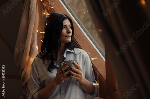 A woman in a shirt is drinking coffee by the unusual window.