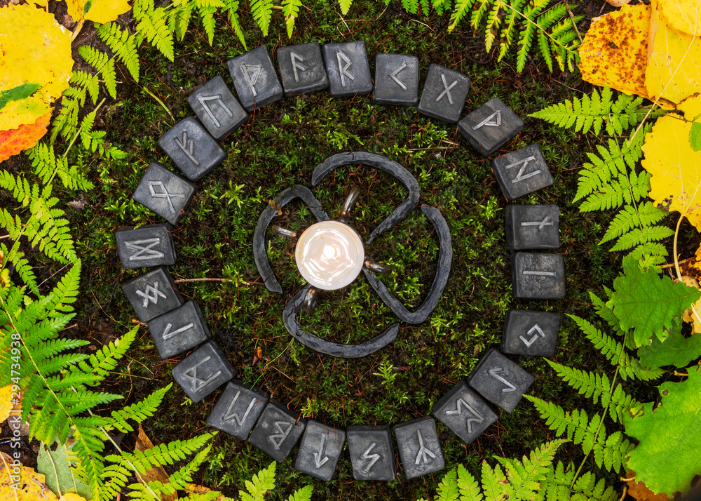 A circle of 24 Scandinavian runes on the background of moss and leaves in the autumn forest. In the center of the circle is a forged candleholder with a candle.