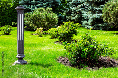 Ground iron lighting lantern mounted on green grass in a park with bushes and pine trees in a well-kept park.