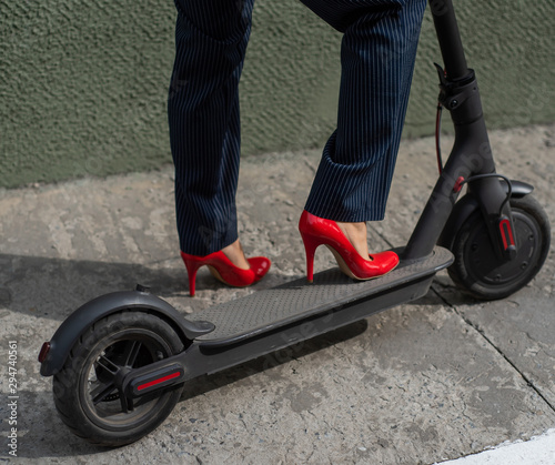 Young woman in formal wear on red hight heels is standing on electrical scooter. Close-up of female legs. A business woman in a trouser suit and red shoes moves around the city on an electric scooter.