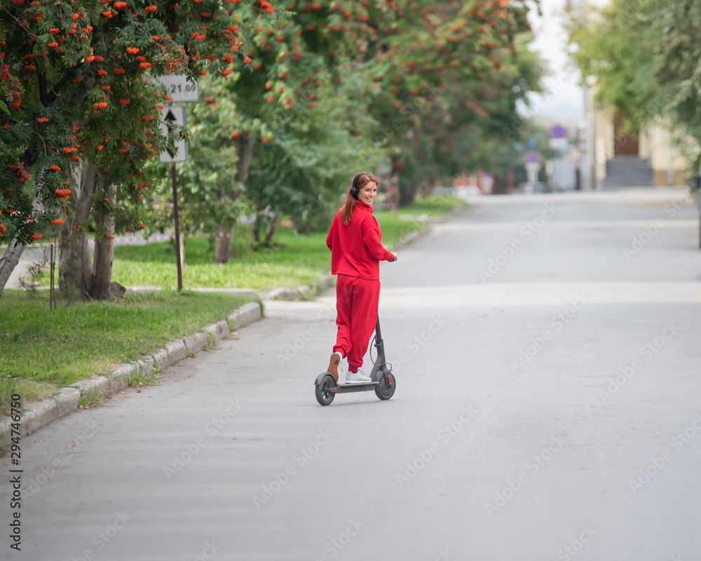 Red-haired girl in a red tracksuit drives an electric scooter. A young woman in oversized clothes rides around the city and listens to music using wireless headphones. View from the back. Red-haired girl in a red tracksuit drives an electric scooter. A young woman in oversized clothes rides around the city and listens to music using wireless headphones. View from the back.
