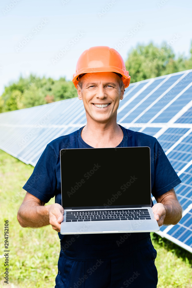 handsome engineer in t-shirt and orange hardhat smiling and holding laptop with copy space handsome engineer in t-shirt and orange hardhat smiling and holding laptop with copy space