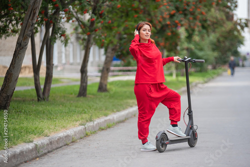 Red-haired girl in a red tracksuit drives an electric scooter. A young woman in oversized clothes rides around the city on modern transport and listens to music using wireless headphones.