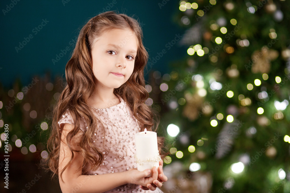 Portrait of acute long-haired little girl in dress on background of  lights.Little girl holding burning candle. Christmas, New Year and birthday celebration concept. Winter holidays. Copy space