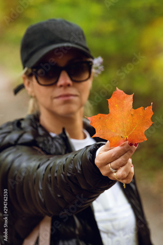 unrecognizable woman in dark hiking jacket, white shirt and black cap holding orange maple leaf in the beginning of autumn in canada., very blurred background, small depth of field